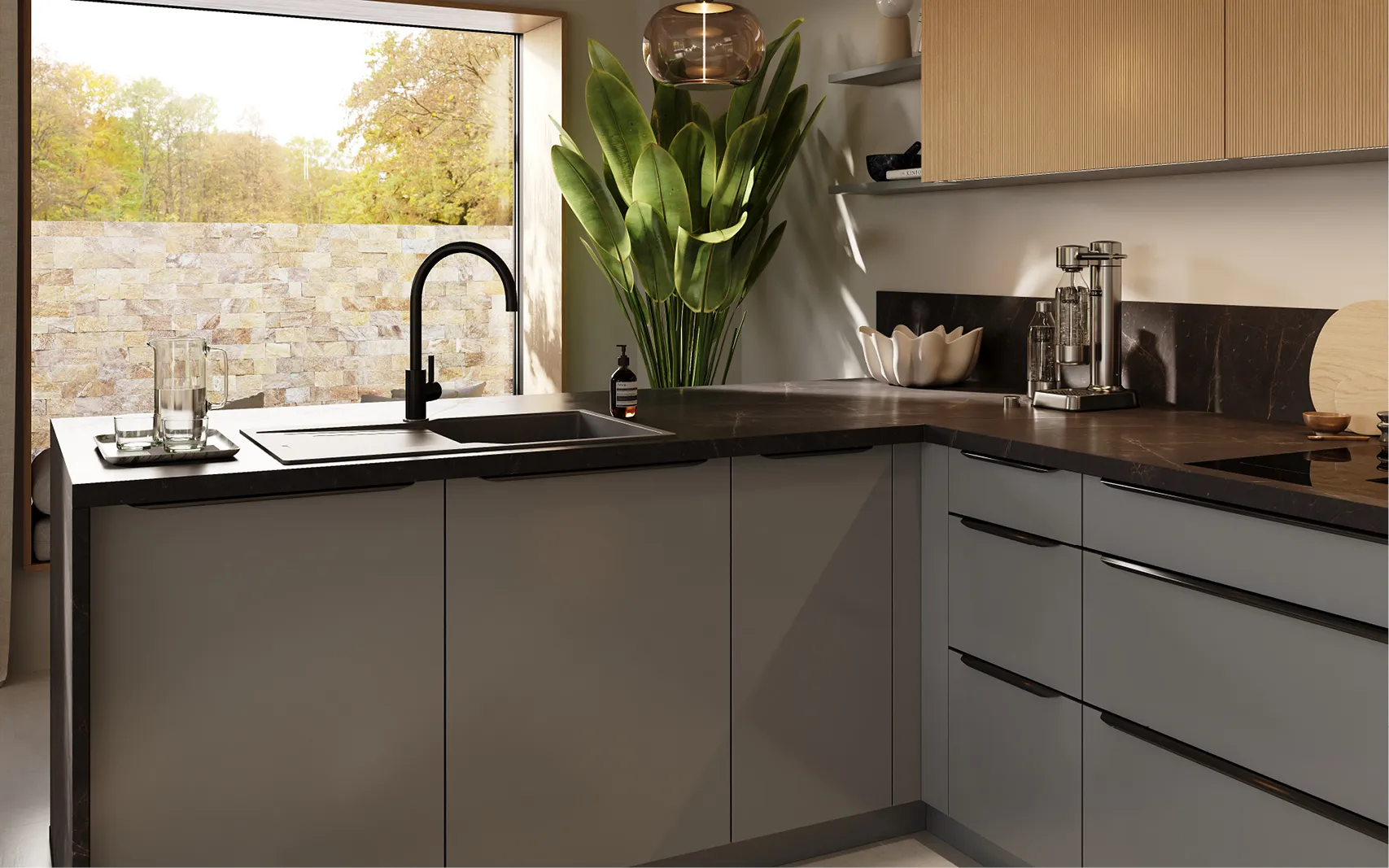 Detailansicht einer modernen L‑Küche: matte graue Fronten, dunkle Stein‑Arbeitsplatte mit Spüle und schwarzem Wasserhahn, große Zimmerpflanze, Fenster mit Natursteinmauer, rechts Schubladen, Induktionsfeld und Wassersprudler.